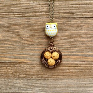 K&ouml;nnte beinhalten: Ein gelber und wei&szlig;er Eulenschmuck h&auml;ngt an einer braunen Kette mit einem Drahtnest, das vier gelbe, gefleckte Eier enth&auml;lt.