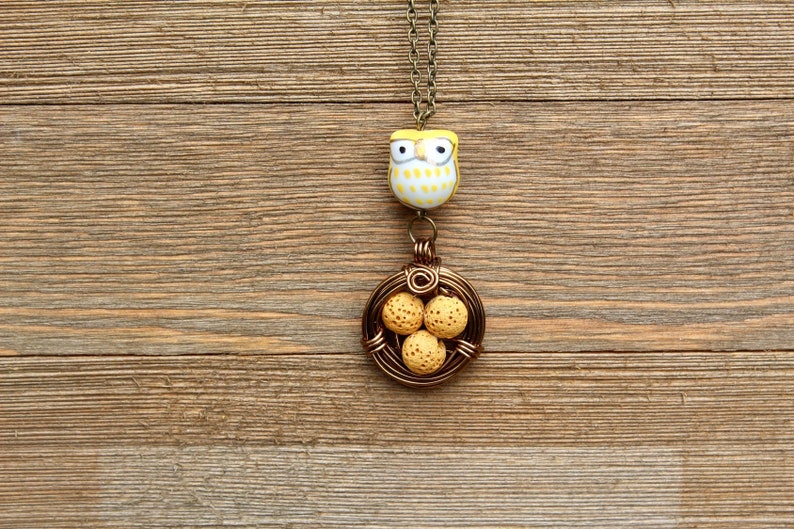 K&ouml;nnte beinhalten: Ein gelber und wei&szlig;er Eulenschmuck h&auml;ngt an einer braunen Kette mit einem Drahtnest, das vier gelbe, gefleckte Eier enth&auml;lt.