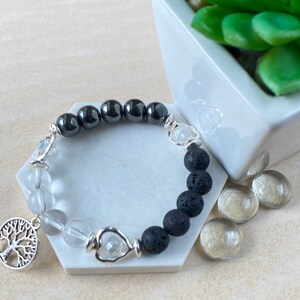 Pode incluir: Um bracelete de contas pretas e brancas com um amuleto de árvore da vida prateado. O bracelete apresenta contas de rocha de lava preta, contas de quartzo transparente e contas de hematita preta.