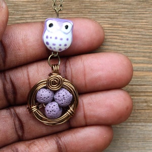 Puede incluir: Collar hecho a mano con un colgante de búho morado y blanco sobre un nido de alambre dorado que contiene tres cuentas de color lavanda. La cadena fina crea una joya caprichosa e inspirada en la naturaleza, ideal para un look único.