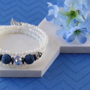 Pode incluir: Um bracelete de contas brancas envolvente com contas de lava azul e cristal. O bracelete está sobre uma superfície hexagonal branca.