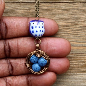Puede incluir: Un amuleto de búho azul y blanco cuelga de una cadena de bronce, con un colgante envuelto en alambre que presenta cuentas de roca de lava azul en un diseño similar a un nido.
