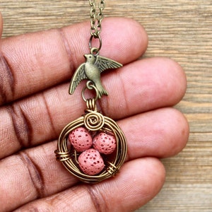 Puede incluir: Un collar de oro con un dije de pájaro y un nido envuelto en alambre con tres cuentas de lava rosa.