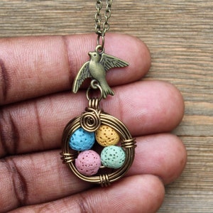 Puede incluir: Un collar de oro con un dije de pájaro y un nido envuelto en alambre con cuatro huevos de roca de lava coloridos. Los huevos están pintados en tonos de azul, amarillo, verde y rosa.
