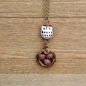 Puede incluir: Un collar con un dije de b&uacute;ho marr&oacute;n y blanco con un colgante de jaula de alambre marr&oacute;n que contiene cuatro cuentas de lava marr&oacute;n.