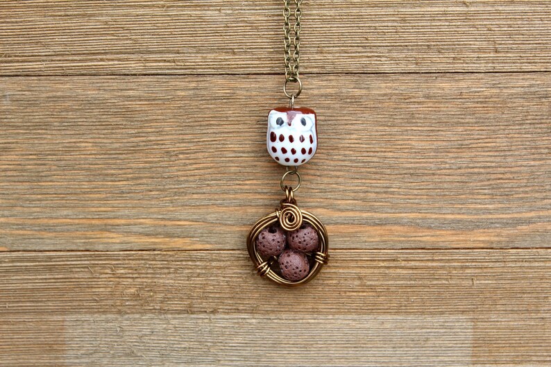 Puede incluir: Un collar con un dije de b&uacute;ho marr&oacute;n y blanco con un colgante de jaula de alambre marr&oacute;n que contiene cuatro cuentas de lava marr&oacute;n.