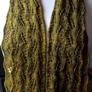 May include: A knitted scarf in shades of olive green and dark brown, displayed on a white mannequin. The scarf features a detailed, openwork pattern, creating a textured and visually interesting design. The scarf is made of soft yarn.