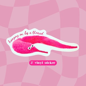 May include: A pink worm-like creature with googly eyes and the text "hanging on by a thread" on a white background. The text "3" vinyl sticker" is below the worm.