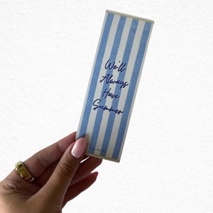 May include: A rectangular bookmark with blue and white vertical stripes. The text "We'll Always Have Summer" is written in blue script. The bookmark is held by a hand with a gold ring.
