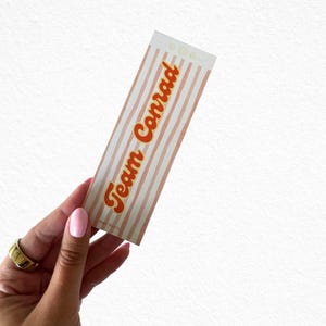 May include: A rectangular sticker with a striped pattern in white and light orange, featuring the text "Team Conrad" in orange script. The sticker is held by a hand with a gold ring and pink nail polish.