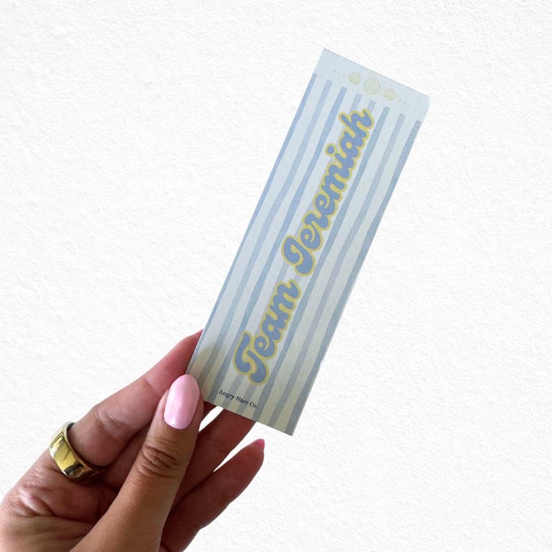 May include: A rectangular bookmark with a light blue and white striped pattern. The text "Team Jeremiah" is written in a yellow, cursive font. The bottom of the bookmark has the text "Angry Hare Co."
