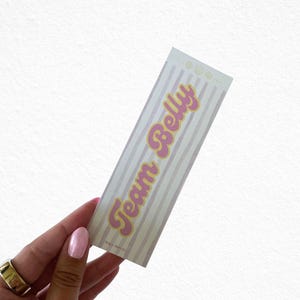 May include: A rectangular sticker with a striped background in light pink and white. The text "Team Belly" is in pink with a yellow outline. The sticker has a retro, playful design.