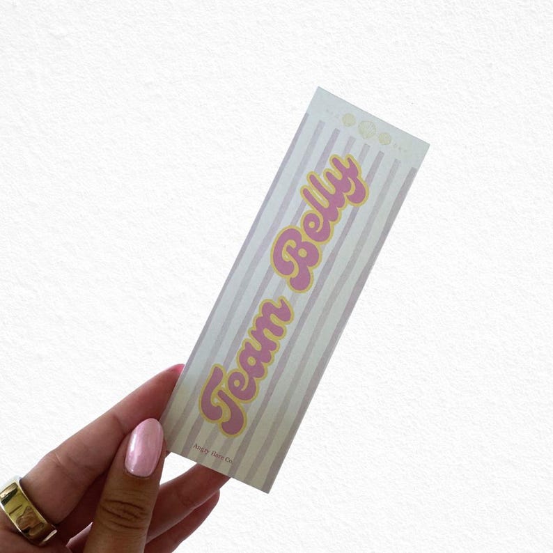 May include: A rectangular sticker with a striped background in light pink and white. The text "Team Belly" is in pink with a yellow outline. The sticker has a retro, playful design.