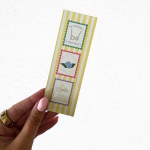 May include: A yellow and white striped bookmark with three postage stamp-style designs. The top stamp reads "THE SUMMER TURNED PRETTY" with a glasses graphic. The middle stamp has a blue flower and reads "IT'S NOT SUMMER WITHOUT YOU". The bottom stamp has a moon and clouds and reads "WE'LL ALWAYS HAVE SUMMER".