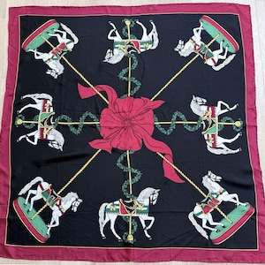 May include: Square black scarf featuring a carousel horse pattern. White horses with red, green, and gold details are set against a black background. A large red bow is in the center, with a burgundy border.