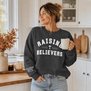 May include: A dark charcoal gray long-sleeve crewneck sweatshirt featuring the white text "RAISING BELIEVERS" with a cross symbol in the center. The right sleeve cuff displays the names "MCKENNA," "MAISIE," and "MILLIE" in white text.