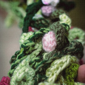May include: A close-up of a crocheted garland with green, pink, and white flowers. The garland is made of yarn and is a handmade craft.