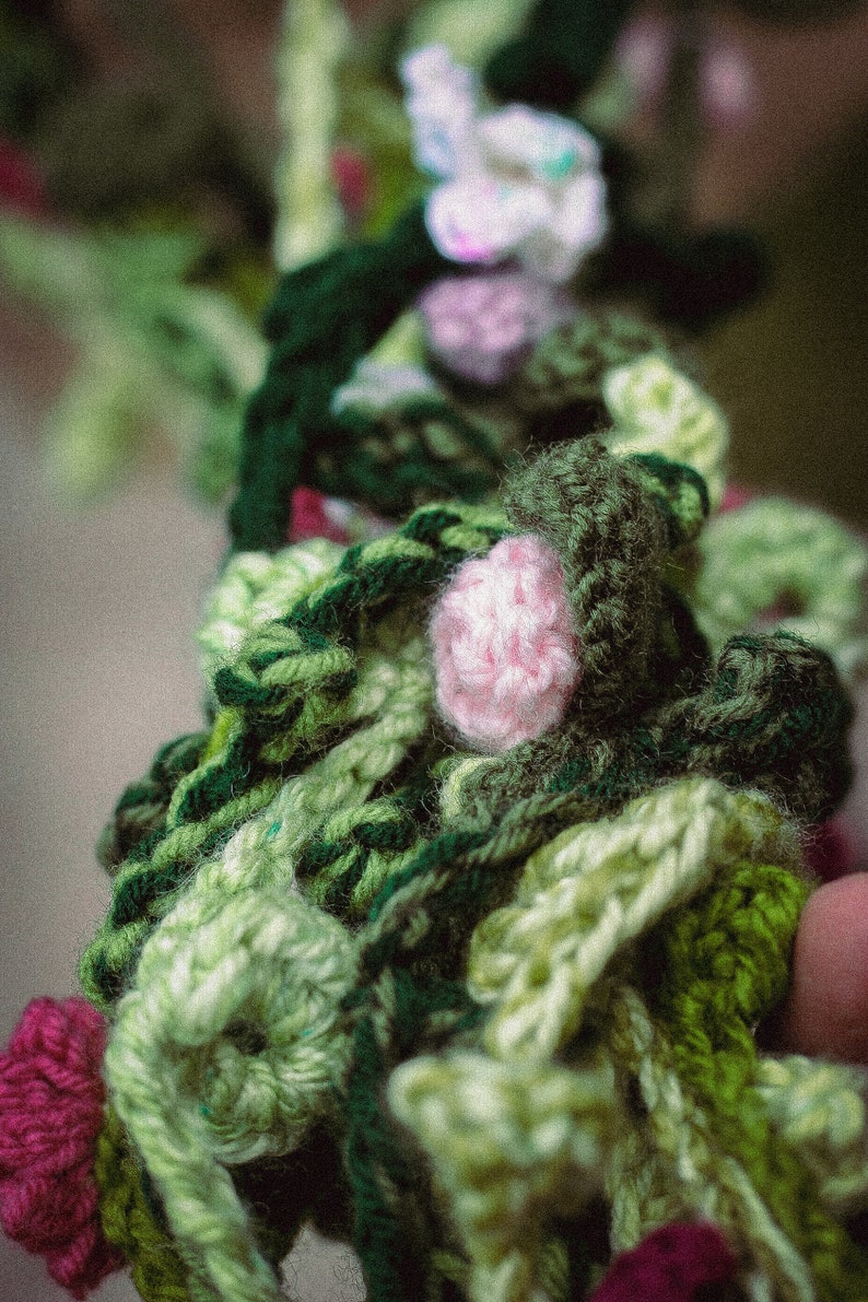 May include: A close-up of a crocheted garland with green, pink, and white flowers. The garland is made of yarn and is a handmade craft.