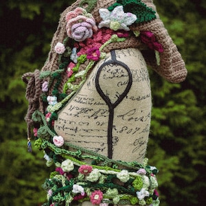 May include: A crocheted shawl with a floral design in shades of pink, green, and white. The shawl is draped over a dress form with a brown background.