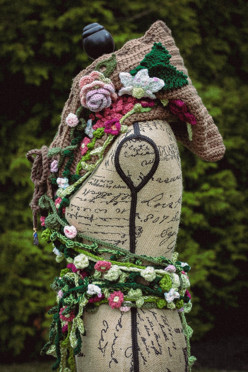 May include: A crocheted shawl with a floral design in shades of pink, green, and white. The shawl is draped over a dress form with a brown background.