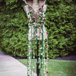 May include: A crocheted hooded top with a floral design in shades of green, white, and pink. The top is displayed on a black stand.