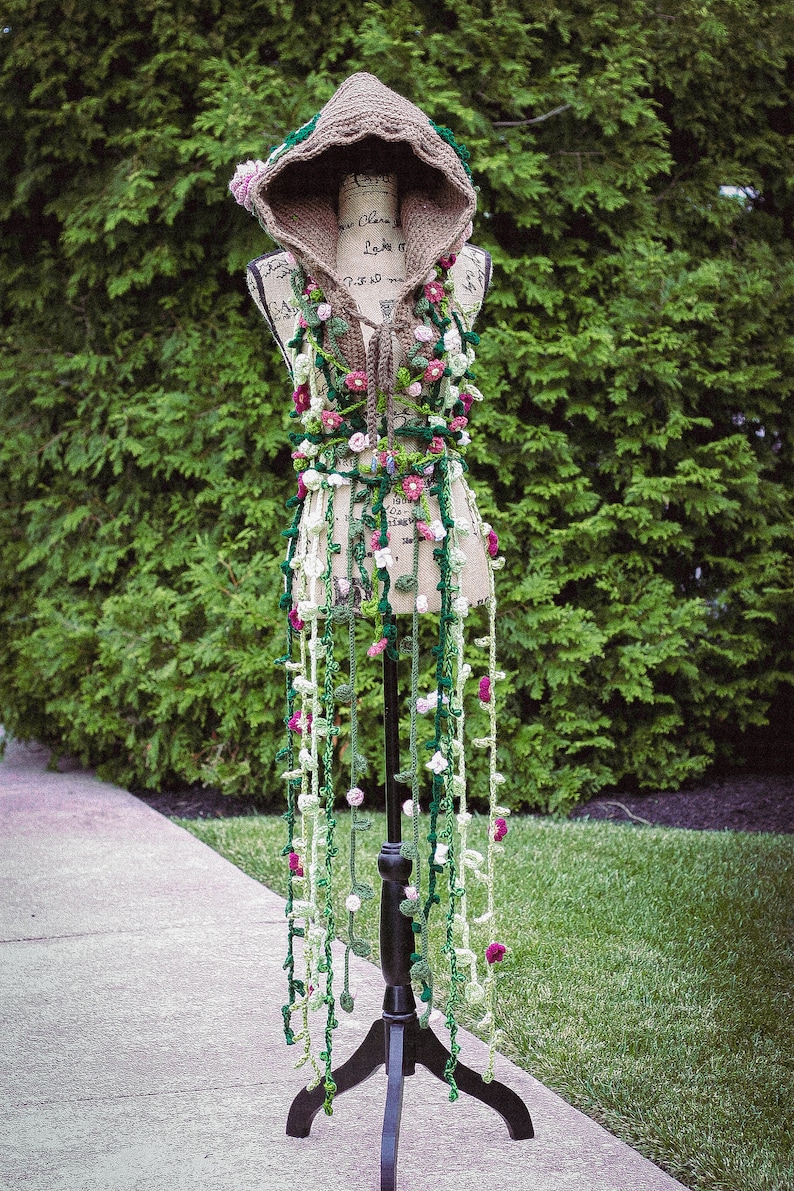 May include: A crocheted hooded top with a floral design in shades of green, white, and pink. The top is displayed on a black stand.