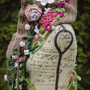 May include: A brown crocheted hooded scarf with floral embellishments. The scarf is decorated with pink, white, and green crocheted flowers and leaves. The scarf is draped over a fabric-covered mannequin.