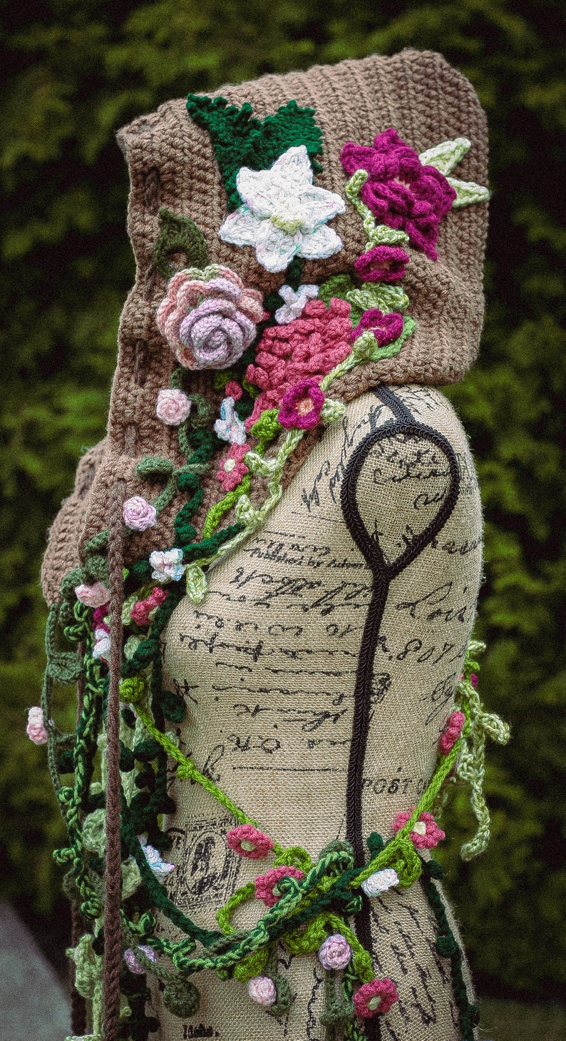 May include: A brown crocheted hooded scarf with floral embellishments. The scarf is decorated with pink, white, and green crocheted flowers and leaves. The scarf is draped over a fabric-covered mannequin.