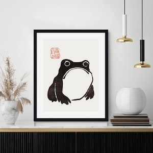 Könnte beinhalten: Minimalistischer Schwarzweißdruck eines Frosches mit einem roten japanischen Stempel im oberen linken Eck. Der Druck ist in einem schwarzen Rahmen gerahmt und auf einem Regal mit anderem Dekor ausgestellt.
