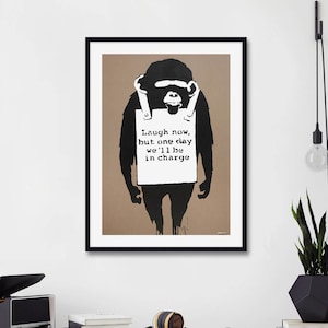 May include: Black and white print of a chimpanzee holding a sign that reads "Laugh now, but one day we'll be in charge". The print is framed in black and hanging on a white wall.