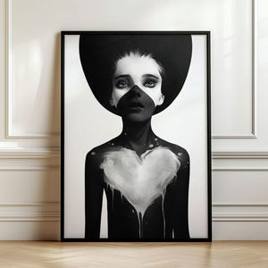 Könnte beinhalten: Ein gerahmter Schwarz-Weiß-Kunstdruck, der das Porträt einer Person mit einem großen kreisförmigen Hut zeigt. Die Figur trägt eine dunkle Gesichtsmaske und einen schwarz bemalten Oberkörper mit einem weißen Herz in der Mitte, das Tropfeffekte aufweist.