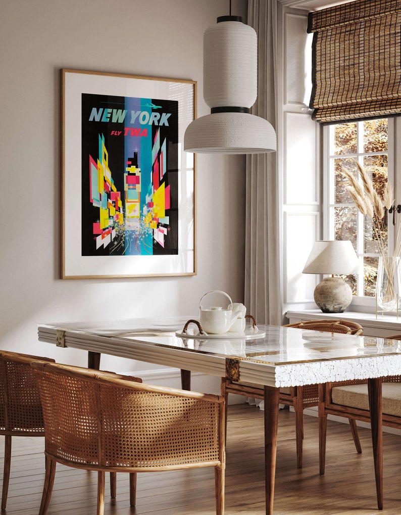 Pu&ograve; includere: Una sala da pranzo con un tavolo bianco con piano in marmo e una lampada a sospensione in ceramica bianca sopra di esso. Ci sono quattro sedie in vimini intorno al tavolo e un poster vintage di New York City sulla parete dietro il tavolo.