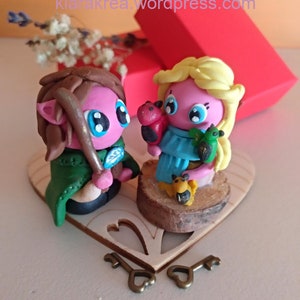 May include: A handmade figurine set of two people, a female with blonde hair and a male with brown hair, holding small animal figurines. The figurines are standing on a heart-shaped wooden base with two keys.