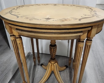 May include: A set of three nesting tables with a light wood finish and intricate floral designs on the top surface. The tables have a rounded oval shape and are supported by slender legs.