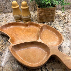 May include: A wooden serving tray with three compartments and a handle. Two yellow salt and pepper shakers with white stripes are behind the tray. A small square woven basket with a green plant is also in the background.