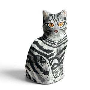 May include: A decorative cat figurine featuring a white base with bold black tabby stripes. The cat has bright orange eyes, a small pink nose, and pointed ears. It is depicted in a seated position with its tail wrapped around its side, showcasing detailed hand-drawn style markings across its entire body.