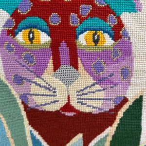 May include: A needlepoint tapestry featuring a stylized leopard face with a red forehead, purple cheeks, and yellow eyes. The leopard is surrounded by green and blue foliage. The design uses vibrant thread colors including teal, lavender, and gold accents on a cream-colored canvas background.