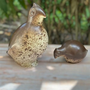 May include: Two ceramic bird figurines with a smooth, glossy finish. The larger bird is upright with a cream-colored body featuring brown speckles. The smaller bird is positioned horizontally, featuring a dark brown head and a speckled cream base.