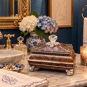 May include: A rectangular decorative box with a distressed bronze and copper finish, featuring a clear faceted crystal knob on the lid and four ornate silver-toned feet. The box has a textured surface and a rope-like trim detail along the lid edge.