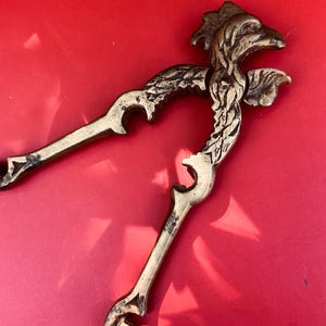 May include: Antique brass nutcracker with a detailed bird design. The nutcracker is open, revealing its lever mechanism. The brass has a tarnished appearance, suggesting age. The background is a solid red.