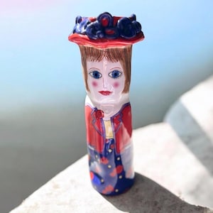 May include: A ceramic vase shaped like a person wearing a red hat with dark blue floral accents, a red jacket, and a blue skirt with red patterns. The figure has brown hair, blue eyes, and red lips, with a cylindrical body and a flared top rim.