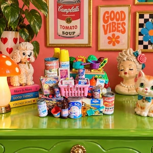 May include: A collection of miniature toy food items and household products, including tiny cans of Campbell's soup, boxes of cereal, and a small pink shopping basket, arranged on a glossy green surface with various decorative figurines nearby.