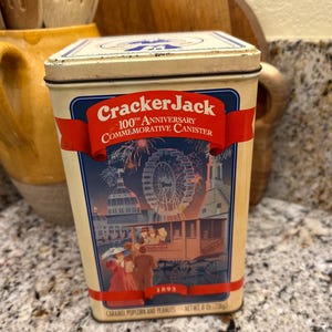 May include: Vintage Cracker Jack 100th Anniversary commemorative canister. The rectangular tin features a cream-colored background with a red banner and blue accents. The design includes an illustration of a Ferris wheel, buildings, and people. The tin contains caramel popcorn and peanuts, net weight 8 oz (226g).