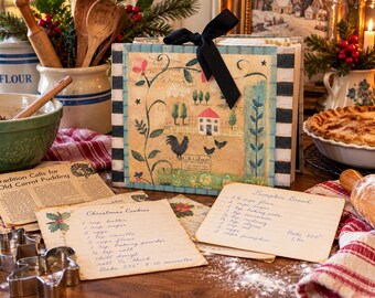 Può includere: Un raccoglitore di ricette quadrato con un nastro nero, caratterizzato da un'illustrazione stampata di una casa, un gallo e polli. Il raccoglitore ha bordi a righe bianche e nere e include schede di ricette scritte a mano per biscotti.