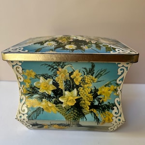 May include: A vintage rectangular tin box with a hinged lid. The box features a floral design with yellow daffodils and other flowers in a woven basket against a light blue background. The lid and edges have a gold trim.
