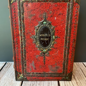May include: A vintage red metal box with a textured pattern and ornate gold and green accents. The box features the words "GOURMETS DELIGHT" in a central oval design. The box has a rectangular shape and is likely used for storage.