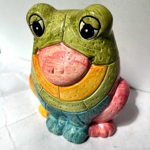 May include: A ceramic frog figurine with a colorful, segmented design. The frog features a green head, a pink mouth, and a body with bands of orange, green, and blue. The eyes are large and black. The figurine is a decorative item.