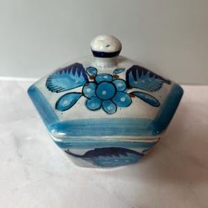May include: A ceramic trinket box with a lid, featuring a blue floral design on a white background. The box has a hexagonal shape with a small knob on top. The edges are outlined in blue, and the overall style is handcrafted.