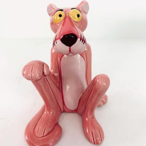 May include: A ceramic Pink Panther figurine in a seated position. The cartoon character is primarily pink with a lighter pink belly and a black nose. The eyes are yellow with black pupils. The figurine has a glossy finish.