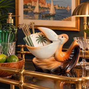 May include: A ceramic pelican figurine with a cream-colored beak and body, featuring brown and black accents on the wings and base. The pelican's open beak holds a stack of square coasters decorated with palm tree illustrations and gold palm tree picks.